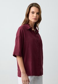Camicia di lino bordeaux con collo a krage, bottoni frontali e maniche corte. Texture liscia e vestibilità rilassata.
