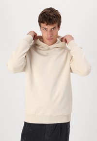 Sudadera beige con diseño de tipo pullover, gran capucha, puños y banda de cintura acanalados. Detalle sutil del logo en el pecho. Textura suave.