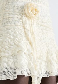 Abito in pizzo color avorio con texture arricciata, caratterizzato da un dettaglio a rosa sul lato e ruffles sovrapposti lungo l'orlo.