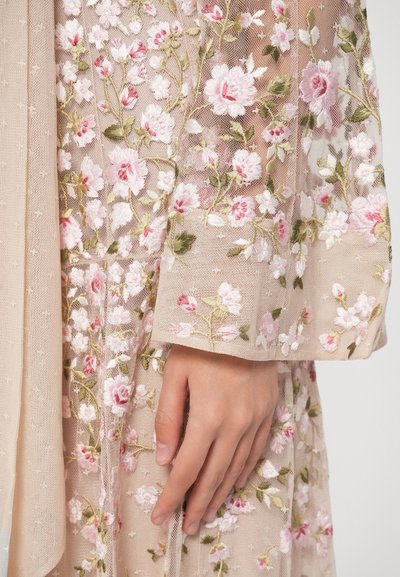 Χέρι resting κατά μήκος ενός μπεζ διαφανούς υφάσματος με περίπλοκη ροζ και λευκή κεντημένη ανθοφορία και πράσινους φύλλους.