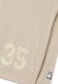 Sudadera beige con una textura suave, que presenta un gran número "35" en blanco desvanecido en el bolsillo y una pequeña etiqueta con la marca en el lado inferior.