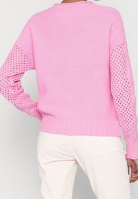 Person wearing a pink knit sweater with openwork sleeve panels and light beige pants, shown from the back against a neutral background.