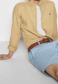 Svetle béžová lněná košile s dlouhým rukávem, bílý tílko, modré šortky a hnědý kožený pásek. Košile má stojící límec.