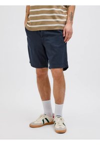 Pantaloni chino blu navy con tasche laterali, vestibilità rilassata e orli rifiniti, indossati con calzettoni bianchi e sneaker crema con strisce verdi.