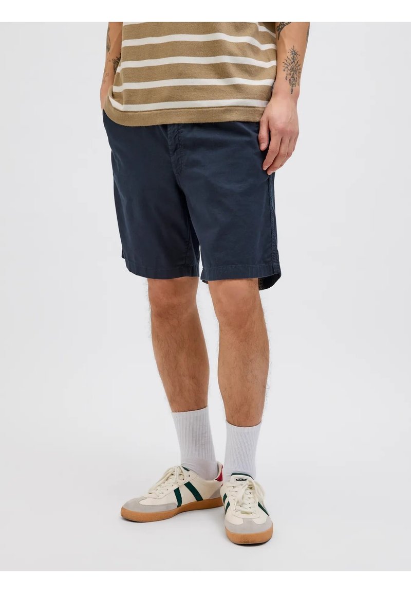 Pantaloni chino blu navy con tasche laterali, vestibilità rilassata e orli rifiniti, indossati con calzettoni bianchi e sneaker crema con strisce verdi.