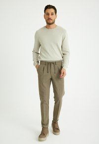 Hellgrauer langärmliger Strickpullover, kombiniert mit sandfarbenen Jogginghosen mit Kordelzug, die ein strukturiertes Muster und eine figurbetonte Silhouette aufweisen, sowie beigen Schuhen.
