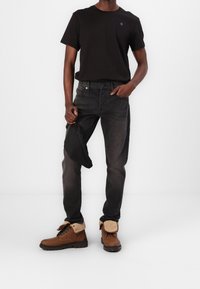 Camiseta de algodón negra, vaqueros de mezclilla gris oscuro y botas marrones con cordones y forro de piel. Sostiene un bolso negro con cremallera. Atuendo simple y casual.