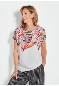Witte blouse met kleurrijke veerpatronen in oranje, paars en zwart. Heeft korte mouwen en een ronde hals. Gecombineerd met gestreepte broek.
