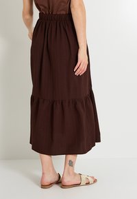 Falda midi marrón en capas con cintura elástica, que presenta una tela texturizada. Llevada con sandalias ligeras y un tatuaje visible en el tobillo.