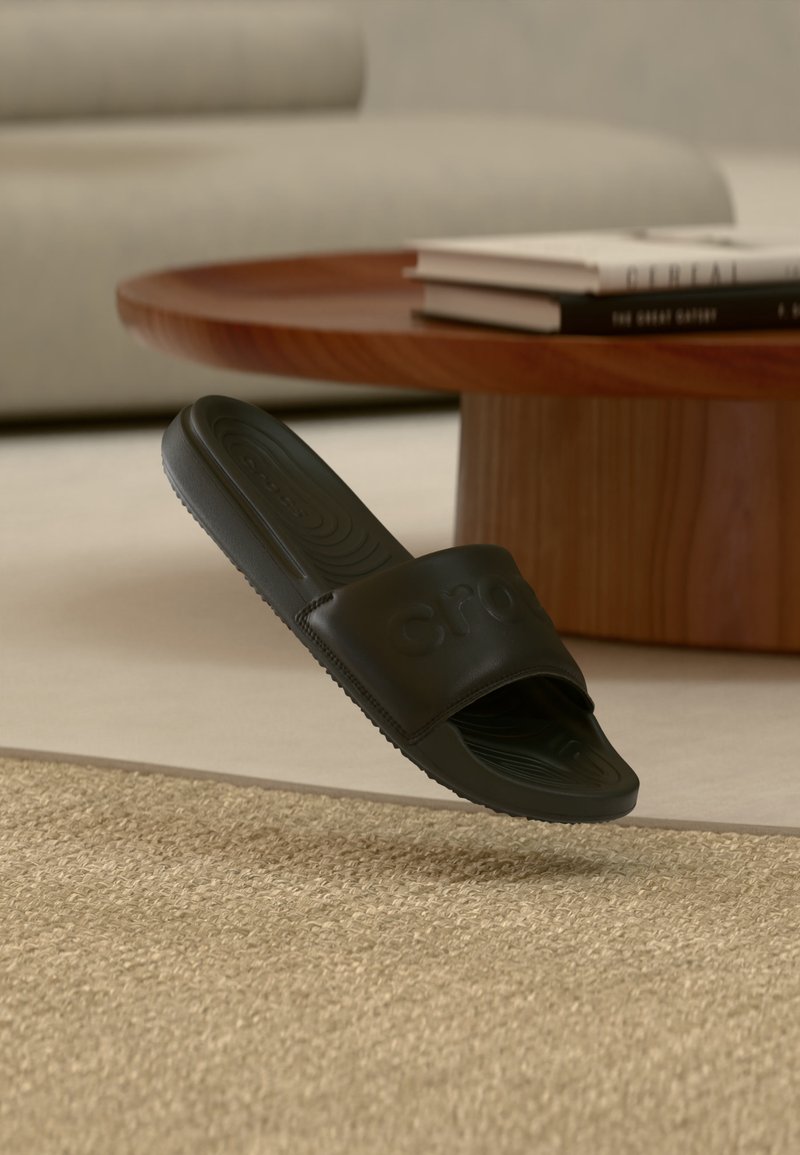 Μαύρο σανδάλι Crocs slide κρεμασμένο πάνω από ένα μπεζ υφασμάτινο χαλί, με ένα στρογγυλό ξύλινο τραπεζάκι καφέ και θολό καναπέ στο παρασκήνιο.