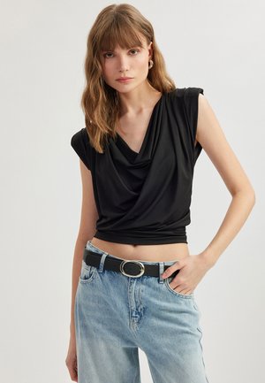Schwarzes ärmelloses Oberteil mit drapiertem Ausschnitt, aus glattem Stoff gefertigt. Kombiniert mit hochgeschnittenen hellblauen Jeans und einem schwarzen Gürtel mit runder Schnalle.