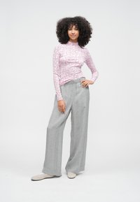 Sieviete stāv ar roku uz gurna, valkājot rozā ziedu raksta garroku topu, pelēkas, platu kāju ziedu raksta bikses un gaišas krāsas apavus uz balta fona.