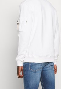 Felpa bianca in cotone con tasca con zip sulla manica, polsini a coste e vestibilità comoda, abbinata a jeans in denim blu.