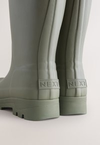 Gros plan de l'arrière de bottes en caoutchouc vert avec des semelles épaisses et « NEXT » embossé sur les languettes de talon.