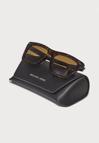 Ochelari de soare din caramel cu lentile întunecate, așezați pe un toc din piele neagră cu textul embosat "MICHAEL KORS."