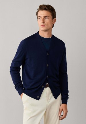 Hombre con un cárdigan azul marino abotonado sobre una camisa oscura y pantalones beige, de pie con una mano en el bolsillo frente a un fondo liso.
