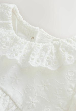 Balts izšūts audums ar viļņotu kakla izgriezumu. Rakstā redzami ziedu motīvi un teksturēti elementi, uzsverot mīkstu, vieglu materiālu.