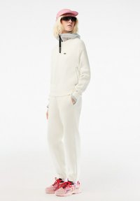Witte sweatshirt van textuurstof, grijze accenten, bijpassende witte joggers, roze pet en roze sneakers met rode veters.