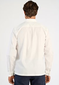 Man met bruin haar, gekleed in een witte lange mouw shirt en donkere broek, gezien van achteren tegen een effen lichte achtergrond.