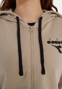 Felpa con cappuccio beige con zip, realizzata in misto cotone, con logo nero, coulisse nere a contrasto e polsini a costine. Texture liscia.