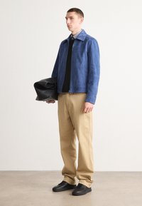 Μπλε σουέτ μπουφάν με κλείσιμο φερμουάρ, σε συνδυασμό με μαύρο πουλόβερ και χακί παντελόνι. Το μοντέλο κρατά μια μαύρη δερμάτινη τσάντα, ενώ στέκεται σε ουδέτερο φόντο.