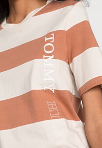 Camiseta a rayas con colores alternos marrón y crema, con "TOMMY HILFIGER" bordado en blanco en el lateral. Mangas cortas.