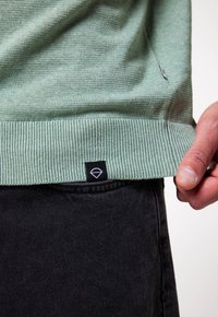 Maglione lavorato a maglia verde chiaro con orlo a costine, caratterizzato da un piccolo etichetta nera con un logo a diamante. Indossato sopra dei jeans in denim neri.