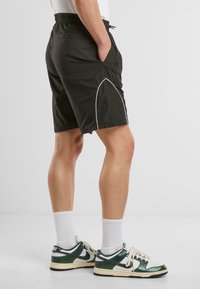 Shorts noirs avec taille élastique et poches latérales, dotés de passepoils blancs le long des coutures latérales. Portés avec des chaussettes blanches et des baskets vertes.