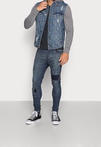 Denimvest med slidte detaljer, grå langærmede skjorte, sort t-shirt, blå ripped jeans og sorte høj-top sneakers med hvide såler.