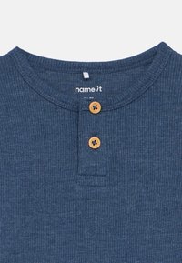 Camisa canelada azul-marinha com decote redondo, apresentando dois botões de madeira no peito e uma pequena etiqueta com a inscrição "name it."