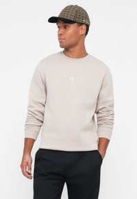 Sudadera gris claro con cuello redondo, que presenta un pequeño texto blanco "II" en el pecho. El modelo lleva una gorra de cuadros con visera oscura.
