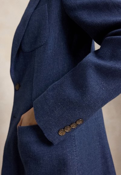 Μανίκι navy blue σακακιού με τέσσερα καφέ κουμπιά και χέρι εν μέρει βουτηγμένο σε τσέπη μπροστά που ταιριάζει.