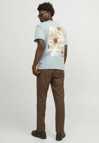 Camiseta de algodón de color azul claro con diseño floral en la espalda, combinada con pantalones marrones y zapatos negros. Modelo usando gafas de sol.
