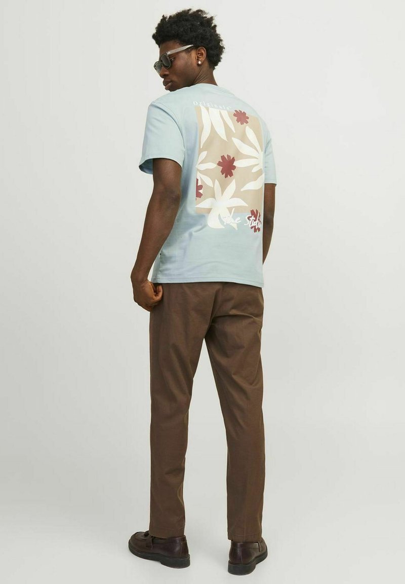 Camiseta de algodón de color azul claro con diseño floral en la espalda, combinada con pantalones marrones y zapatos negros. Modelo usando gafas de sol.