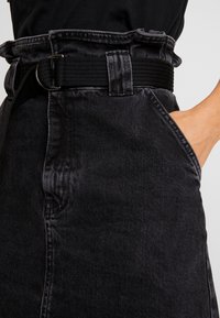 Person som bär högmidjade svarta denimjeans med ett svart tygbälte, höger hand placerad i framfickan.