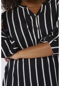 Blouse à rayures noires et blanches avec un col boutonné, des manches longues et une coupe décontractée, mettant en valeur un tissu lisse et des poignets retroussés.