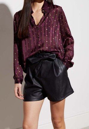 Burgunderfarbene Seidenbluse mit vertikalen Streifen und kleinen goldenen Punkten, kombiniert mit hochgeschnittenen schwarzen Leder-Shorts mit einer Schleifenapplikation.