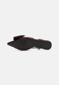 Scarpa con tacco in vernice rossa, punta affilata, cinturino alla caviglia e suola nera texturizzata. Presenta un dettaglio a fiocco nella parte anteriore.