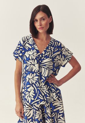 Kobieta o krótkich brązowych włosach, ubrana w niebieską i białą bluzkę z krótkim rękawem w tropikalny liściasty wzór oraz pasującą spódnicę, stojąca z ręką na biodrze.