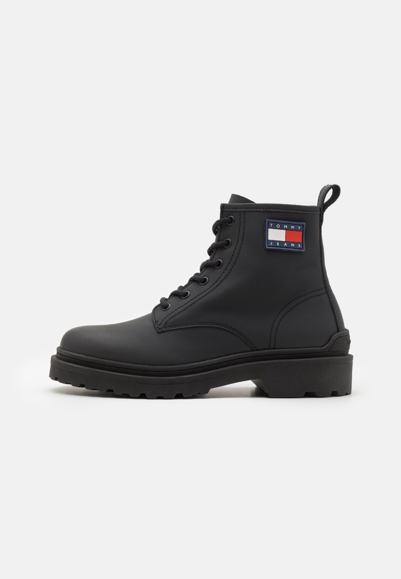 Bottine noire à lacets avec semelle épaisse et languette, présentant un patch logo rectangulaire Tommy Jeans sur le côté extérieur.