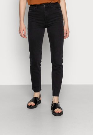 Person iført sorte højttaljede skinny jeans og sorte åbne sandaler, stående mod en lys grå baggrund.