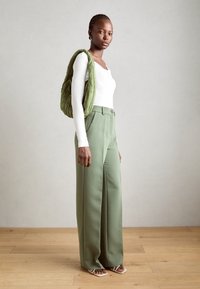 Green wide-leg trousers with a high waist, paired with a white long-sleeve top. A green quilted backpack is worn over one shoulder.
