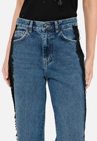 Jeans in denim blu con dettagli in pizzo nero sfrangiato lungo i lati, dotati di vita alta e design classico a cinque tasche.