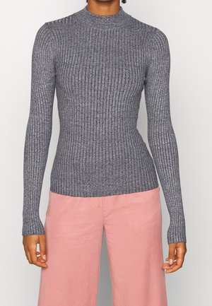 Kvinde iført en tætsiddende, langærmet, ribstrikket grå sweater kombineret med højtaljede, vidde bukser i rosa.