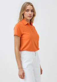 Pomarańczowa koszulka polo z krótkim rękawem, zapinana na guziki, wykonana z bawełny, zestawiona z białymi spodniami o wysokim stanie. Gładka tekstura, luźny krój.