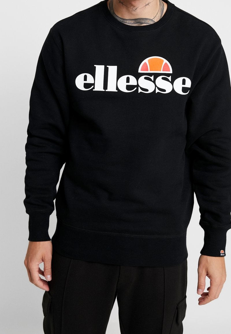 Homme portant un sweat-shirt ellesse noir avec logo blanc et orange, associé à un pantalon noir, sur fond gris uni.