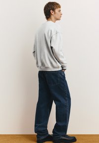 Grauer Sweatshirt mit fallenden Schultern, kombiniert mit lockeren dunkelblauen Jeans. Das Outfit wird durch schwarze Stiefel ergänzt und betont einen lässigen, entspannten Stil.