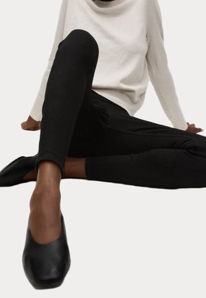 Persona sentada en el suelo que lleva pantalones ajustados negros, zapatos planos negros y un suéter de manga larga beige claro con cuello vuelto.