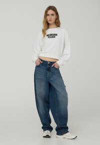 Felpa bianca cropped con testo nero "NO DRAMA PLEASE" abbinata a jeans blu denim larghi. Il modello sta in piedi con le mani in tasca, indossando sneakers bianche.