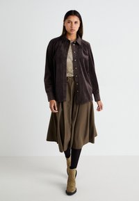 Bruine corduroy overhemd met een kraag, knopen aan de voorkant, gedragen over een lichtgrijze top en een olijfgroene midi-jurk, gecombineerd met tan enkellaarsjes.